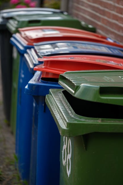 A row of large wheelie bins positioned along a pavement beside a brick wall, with the bins arranged in an alternating pattern of blue, green, and red colours. The bins are made of plastic with texture