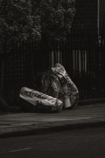 Two large black plastic rubbish bags, visibly crumpled and loosely tied at the top, are placed on a sidewalk curb in front of a wooden fence with vertical slats. The bags appear to contain mixed waste