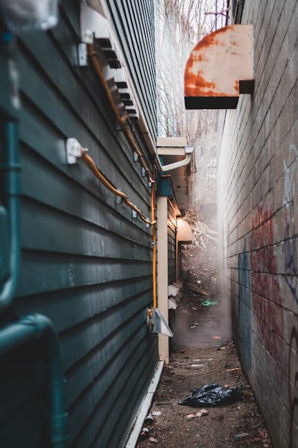 A dirty, weathered, and stained beige fabric trash bag rests on a concrete pavement in a narrow urban street, leaning against a rough, graffitied brick wall. The bag appears to be filled with refuse a